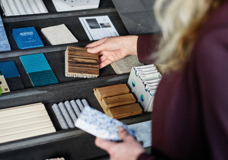 Person betrachtet verschiedene nachhaltige Materialproben, darunter Holz-, Stein- und Texturplatten, die in einem Regal ausgestellt sind.